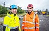 To anleggsarbeidere i synlige verneklær står på en anleggsvei med maskiner i bakgrunnen.